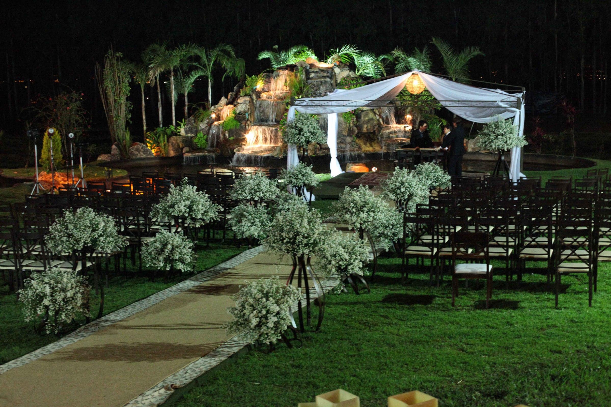 Espaço de cerimônia no jardim à noite, com pergolado e vegetação iluminados e cadeiras alinhadas