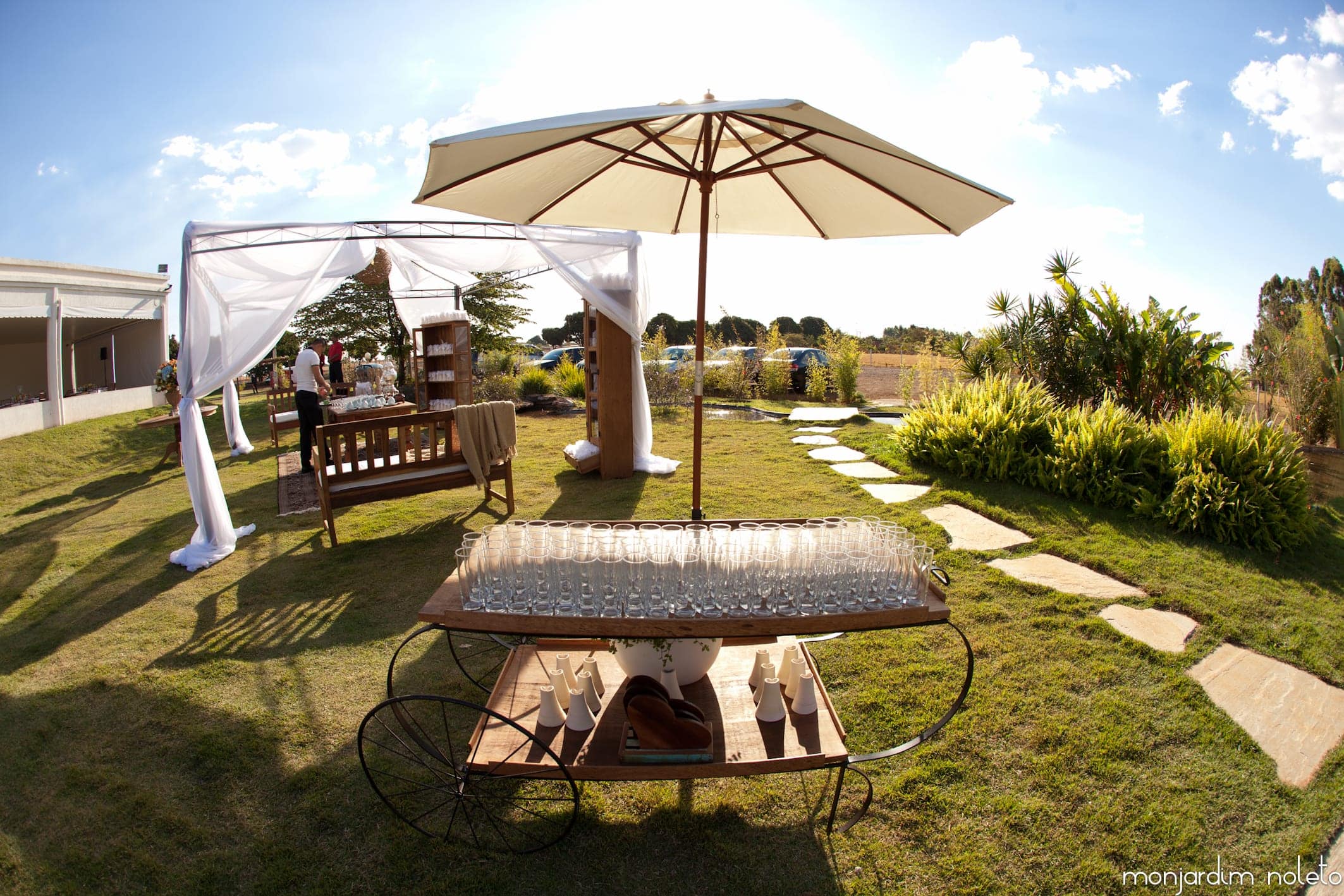 Lounge externo com guarda-sol, banco e mesa de apoio sobre o gramado, durante o dia