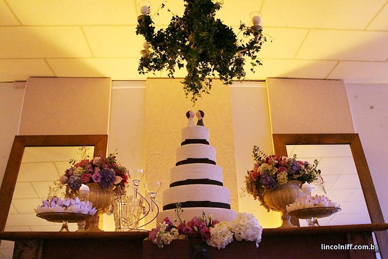 Mesa do bolo montada no salão, com arranjos de flores dos dois lados