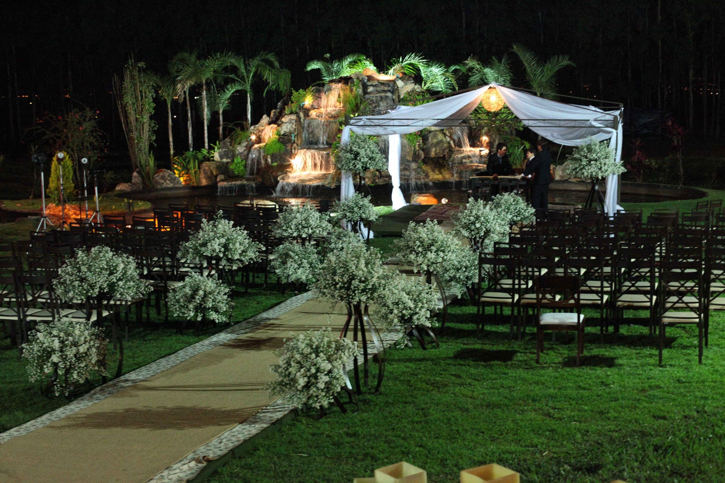 Espaço de cerimônia no jardim à noite, com pergolado e vegetação iluminados e cadeiras alinhadas